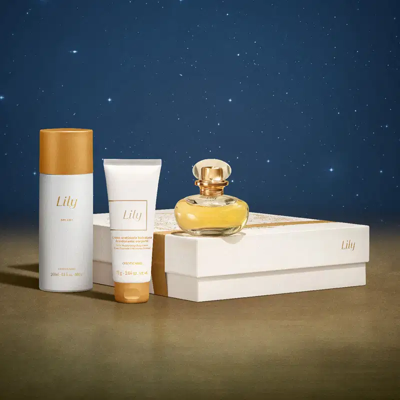 Imagem do Kit Presente Natal Lily com um vidro de desodorante colônia, um de body splash, uma bisnaga de creme hidratante corporal e uma caixa presenteável rosa-claro com estampa floral em cenário com fundo azul-escuro e superfície marrom.