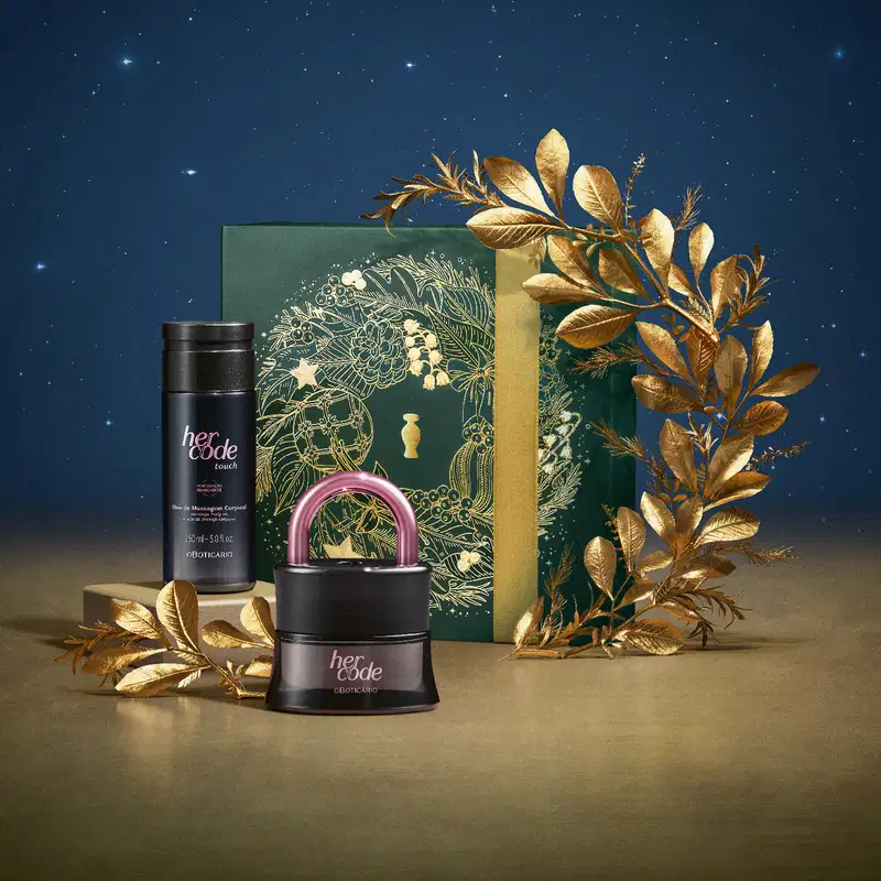 Imagem do Kit Presente Natal Her Code Touch com um vidro de desodorante colônia, um frasco de óleo de massagem corporal e uma caixa verde presenteável com estampa natalina em cenário com flores douradas, superfície marrom e fundo azul.