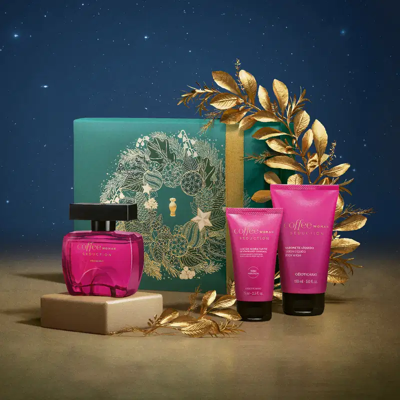 Imagem do Kit Presente Natal Coffee Woman Seduction onde o frasco de desodorante colônia está sobre um pedestal bege. A a caixa de presente e as duas bisnagas de itens de corpo e banho estão sobre uma superfície marrom em um cenário com fundo azul.