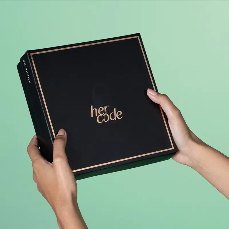 Imagem de caixa preta com texto dourado Her Code sobre fundo verde claro, segurada por mãos femininas.