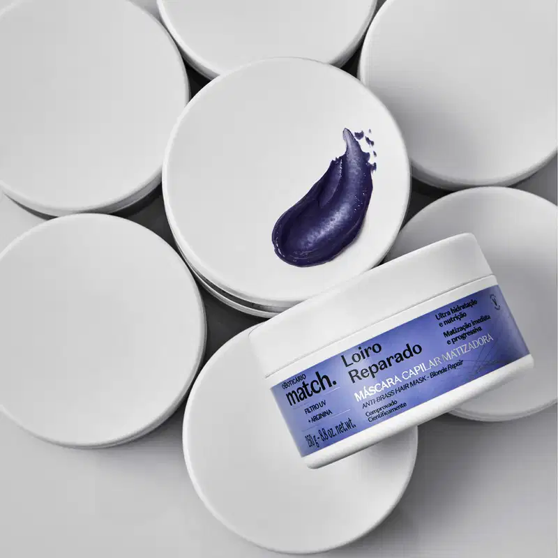 Imagem de vários potes brancos de máscara capilar matizadora da marca Match, com um pouco de creme azul escuro em um deles, sobre uma superfície clara.