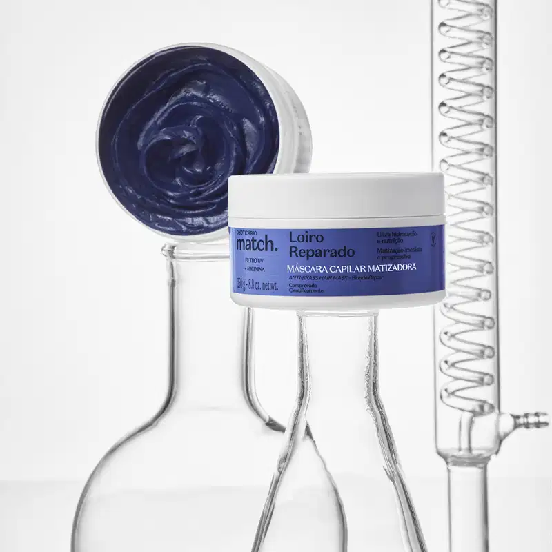 Imagem de um pote branco com rótulo azul da marca Match contendo máscara capilar reparadora, sobre superfície clara com frascos de vidro laboratoriais ao redor. Também mostra a embalagem aberta apresentando um creme azul escuro.