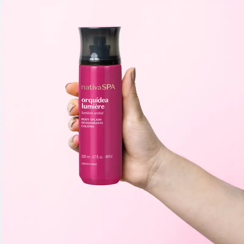 Frasco cilíndrico de item de perfumaria da marca Nativa SPA Orquídea Lumière, na cor rosa vibrante, sendo segurado por uma mão feminina contra um fundo rosa claro.