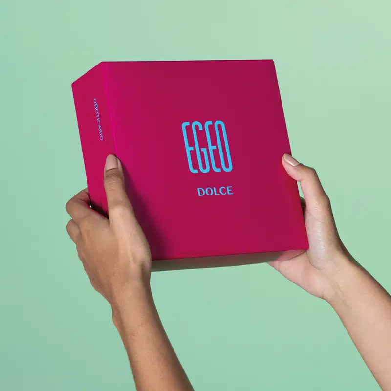 Imagem de uma mão segurando uma caixa rosa vibrante com a marca Egeo Dolce em azul, exibida sobre fundo verde claro, em ambiente minimalista e monocromático.