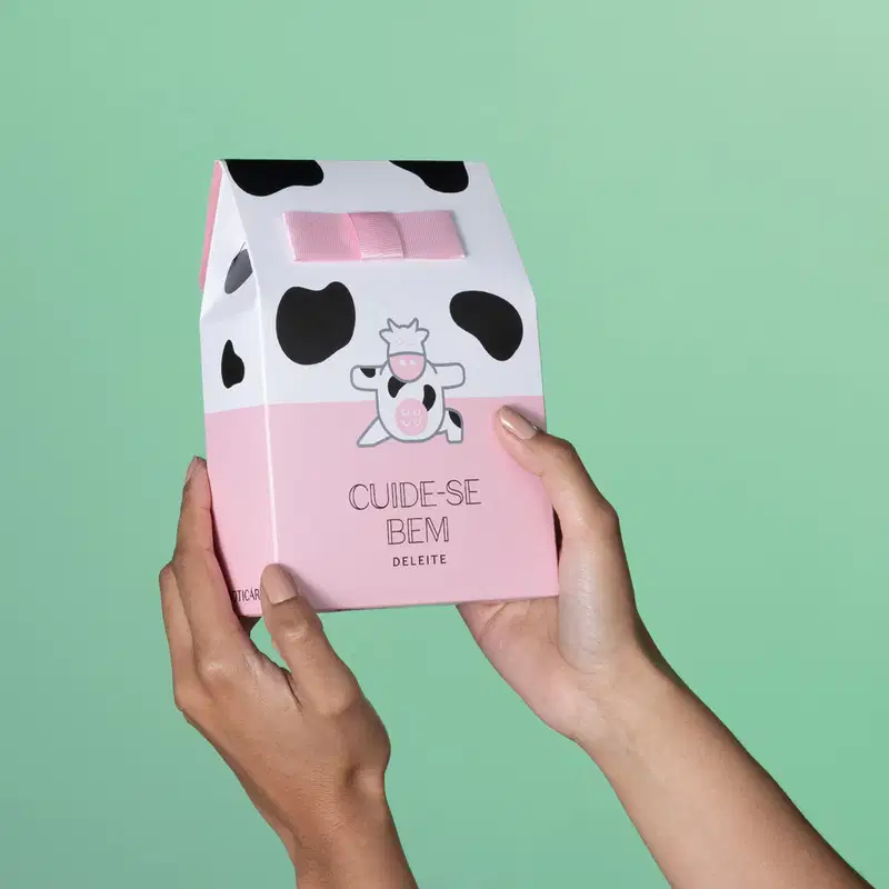 Imagem de uma mão segurando uma sacola presenteável branca e rosa com manchas pretas e ilustração de vaquinha da marca Cuide-se Bem Deleite em fundo verde.