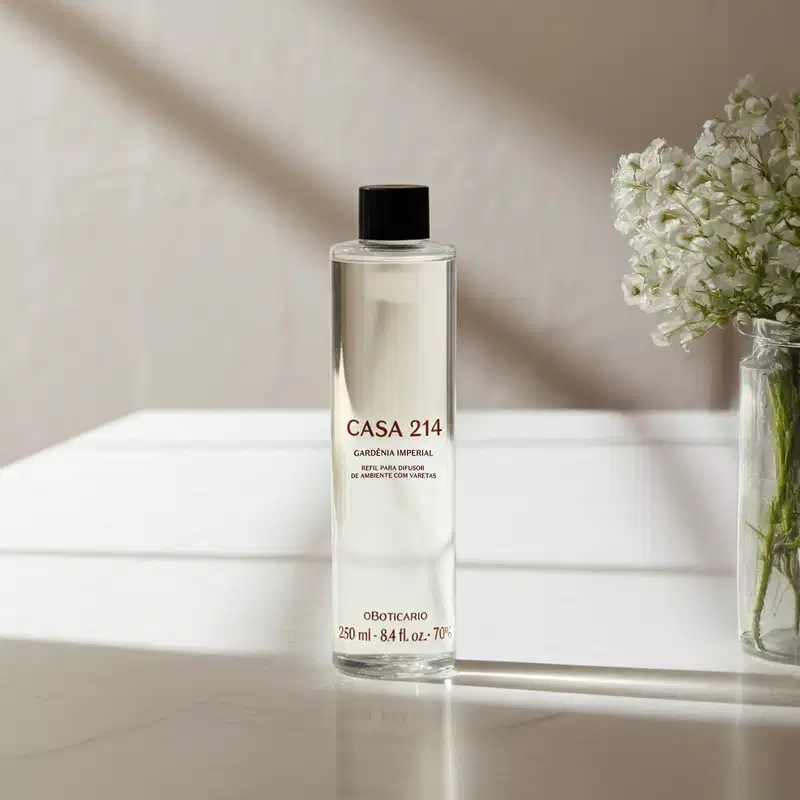 Imagem de um frasco refil com a fragrância Gardênia Imperial para difusor de ambiente da marca Casa 214 em embalagem transparente e tampa preta, posicionado sobre superfície clara com vaso de flores brancas ao fundo.