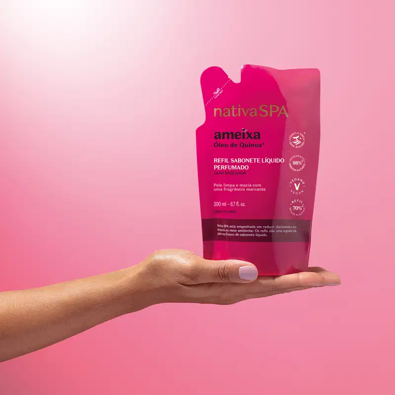 Imagem de uma mão segurando um refil de sabonete líquido perfumado da marca Nativa SPA Ameixa em sachê na cor rosa vibrante e letras pretas e brancas, em fundo rosa.