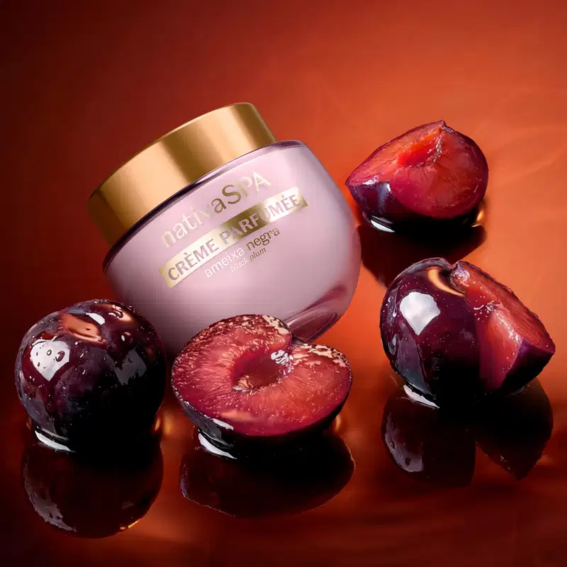 Pote arredondado de creme perfumado Nativa SPA Ameixa Negra, rodeado por ameixas negras cortadas em diversos formatos sobre superfície laranja-avermelhado brilhante que reflete os itens sobre ela.