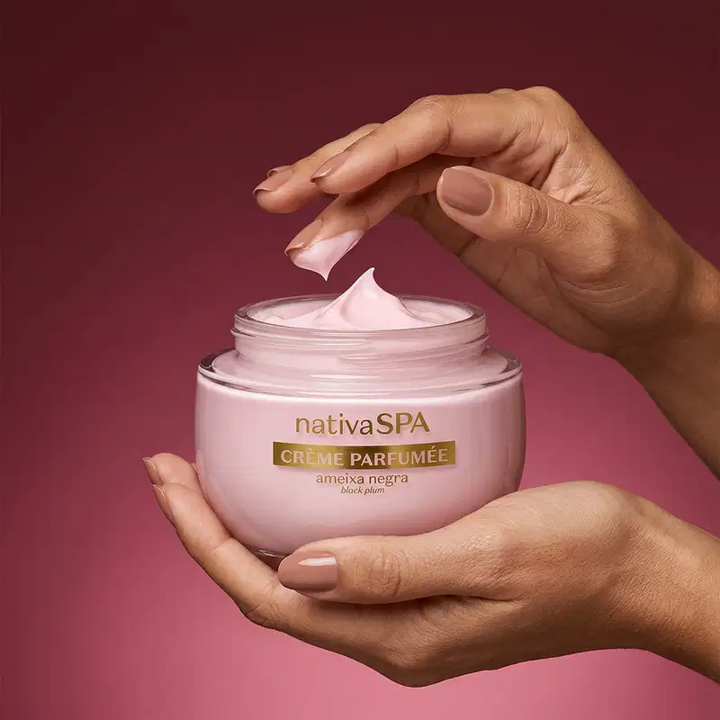 Pote aberto de creme perfumado da marca Nativa SPA Ameixa Negra repousa em mão esquerda feminina enquanto a mão direita repousa acima da embalagem aberta com um pouco de creme na ponta do dedo médio em fundo rosa escuro.