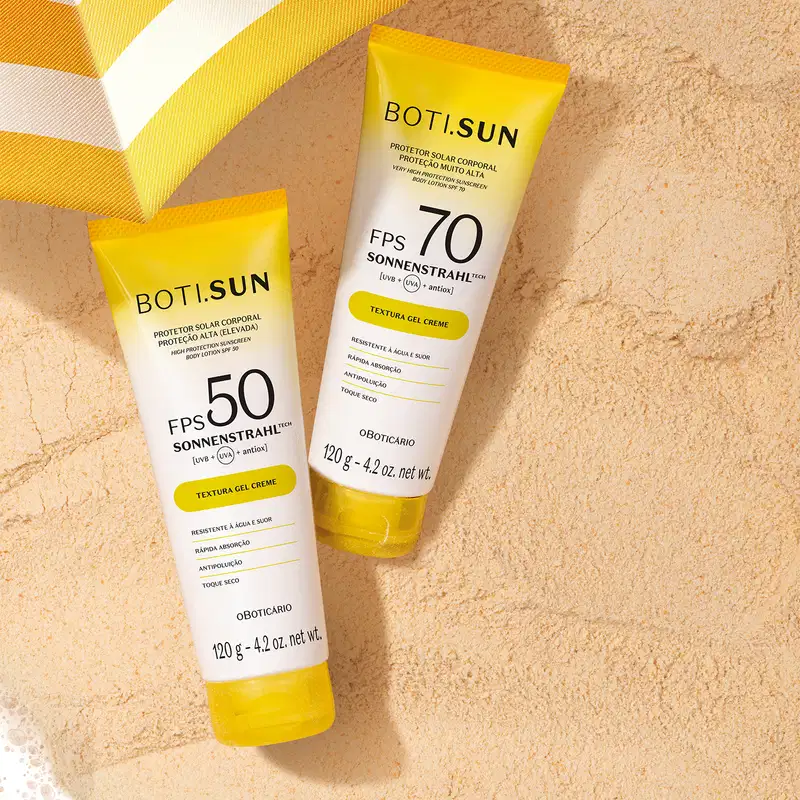 Dois tubos de protetor solar da marca Boti Sun, com FPS 50 e 70, sobre superfície bege simulando areia e parte de um guarda-sol amarelo e branco.