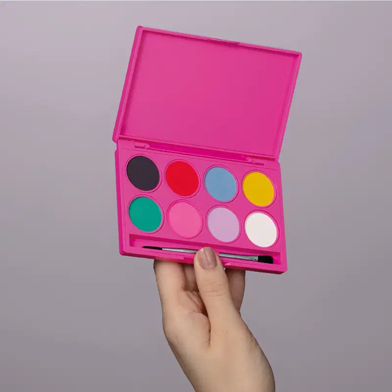 Paleta de maquiagem multifuncional com embalagem rosa e 8 tons vibrantes de maquiagem, espelho na tampa e pincel aplicador sendo seguro por mão feminina em fundo cinza.