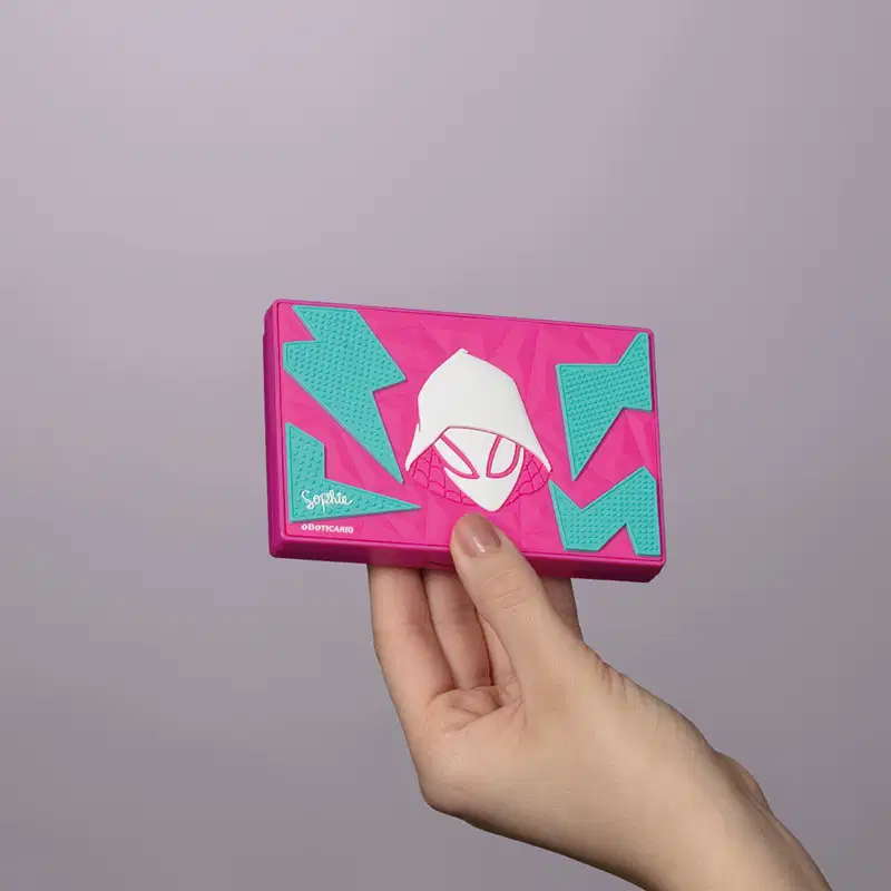 Estojo de maquiagem rosa e azul da marca Sophie Spider-Gwen com logotipo da personagem ilustrado em branco com elementos geométricos azuis ao redor, sendo segurada por mão feminina em fundo cinza.