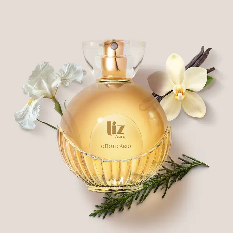 Frasco de desodorante colônia da marca Liz Aura em recipiente de vidro com líquido dourado, rodeado por flores brancas e folhagens verdes sobre fundo bege claro.
