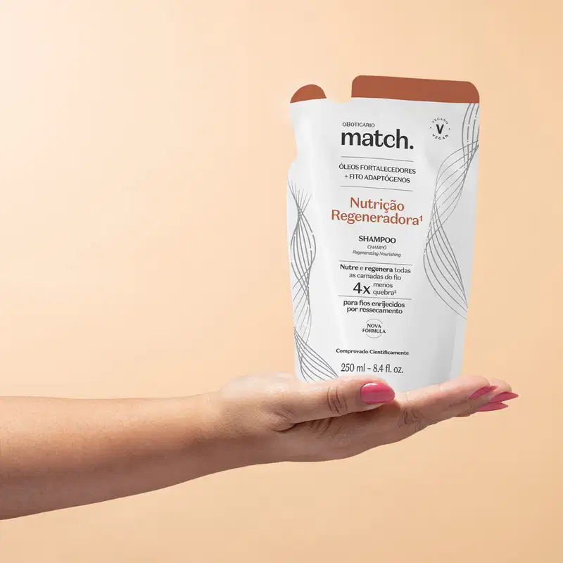Imagem de uma mão feminina segurando um refil em forma de sachê na cor branca com detalhes laranja e letras pretas contendo shampoo da marca Match. Nutrição Regeneradora. O fundo da imagem é rosa.