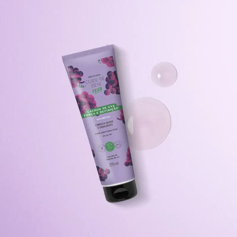 Imagem de um tubo de shampoo da marca Cuide-se bem Feira Cachos de Uva em embalagem roxa com ilustrações de uvas, sobre superfície lilás clara com duas gotas do produto, com textura gelatinosa e brilhante, ao lado.