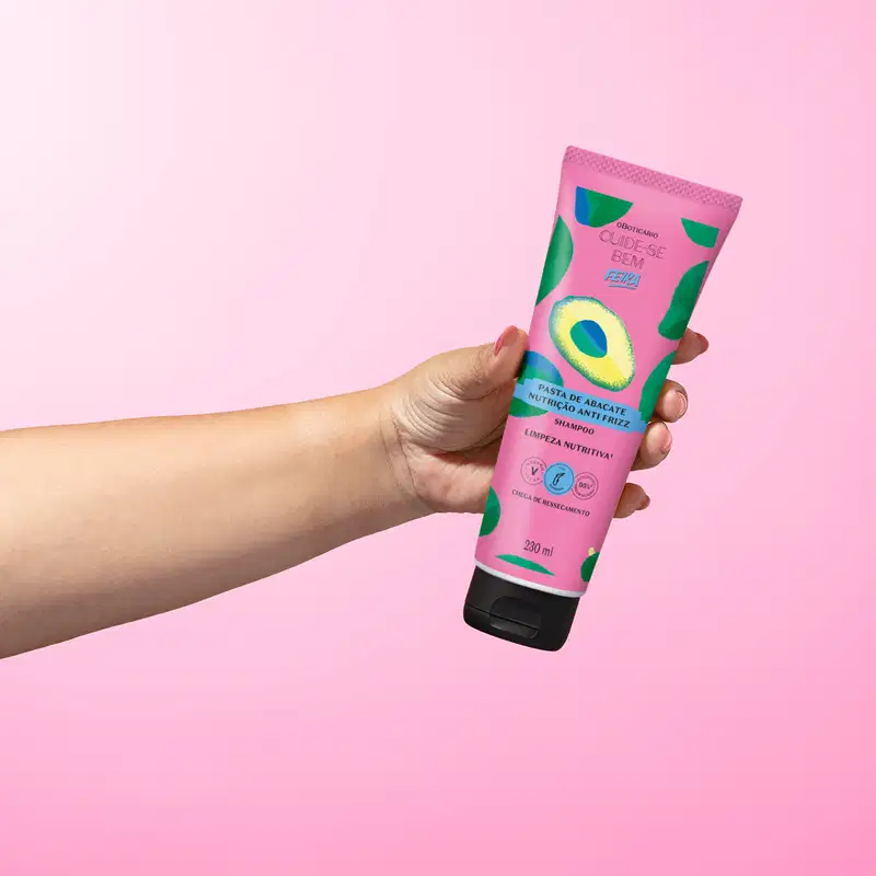 Imagem de um tubo roxo com ilustrações de abacate de shampoo da marca Cuide-se Bem Feira Pasta de Abacate, seguro por mão em fundo rosa.
