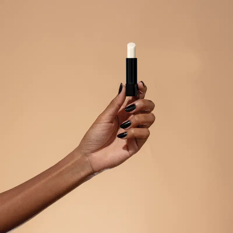 Mão segurando um balm labial noturno da marca Make B. em fundo bege, com unhas pintadas de preto, em um ambiente minimalista e elegante.