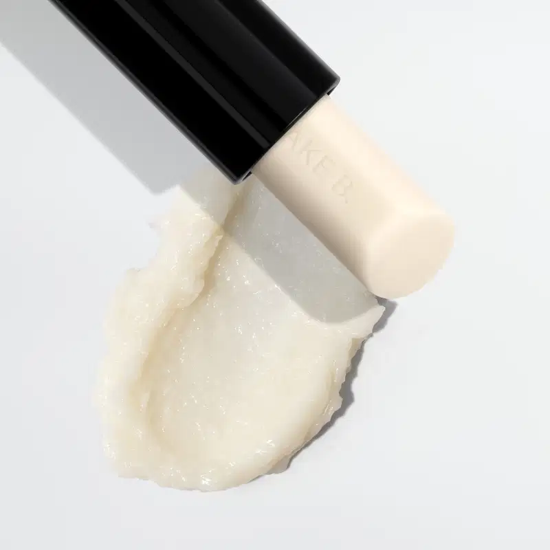 Frasco preto de Balm Labial Noturno Make B. Lip Chronology Night Peeling., contendo produto de textura cremosa e brilhante em tom bege claro.