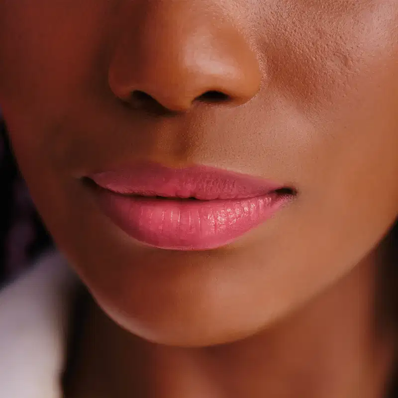 Parte do rosto feminino evidenciando os lábios rosados e brilhantes, sugerindo que a modelo esteja usando o balm labial rosa da marca Make B. Lip Chronology Color Repair.