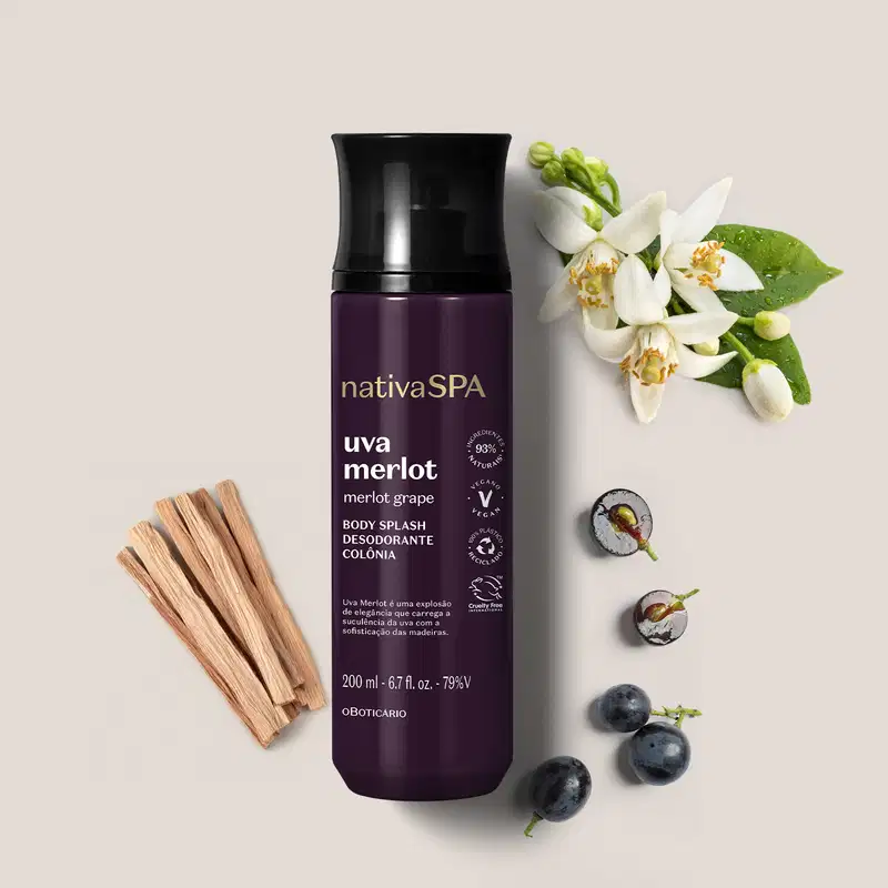 Ao lado do frasco roxo e preto de body splash Nativa SPA Uva Merlot há alguns ingredientes naturais relacionados à fragrância, como uvas e flores brancas. O fundo é lilás bem claro.