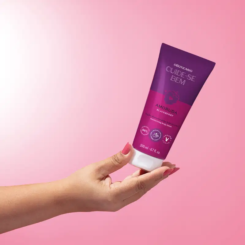 Mão feminina segurando u tubo roxo e branco com ilustração de amora de loção desodorante corporal Cuide-se Bem Amoruda sobre fundo com degradê de branco e lilás.