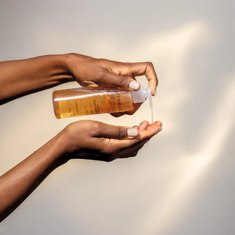 Imagem de duas mãos femininas, uma delas segura o frasco transparente do gel de limpeza Botik Vitamina C na posição horizontal e joga uma quantidade do produto na outra mão. O fundo é cinza claro com leve sombreado.