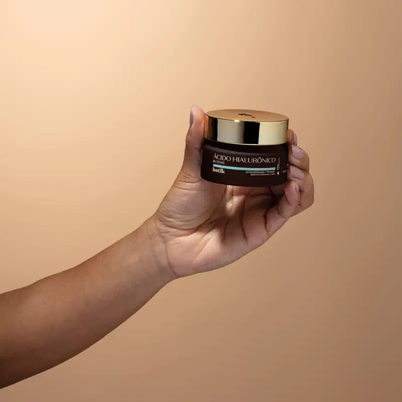 Imagem de recipiente de creme facial com rótulo da marca Botik, sendo segurado por uma mão em fundo bege claro. O produto tem a cor vinho com tampa dourada.