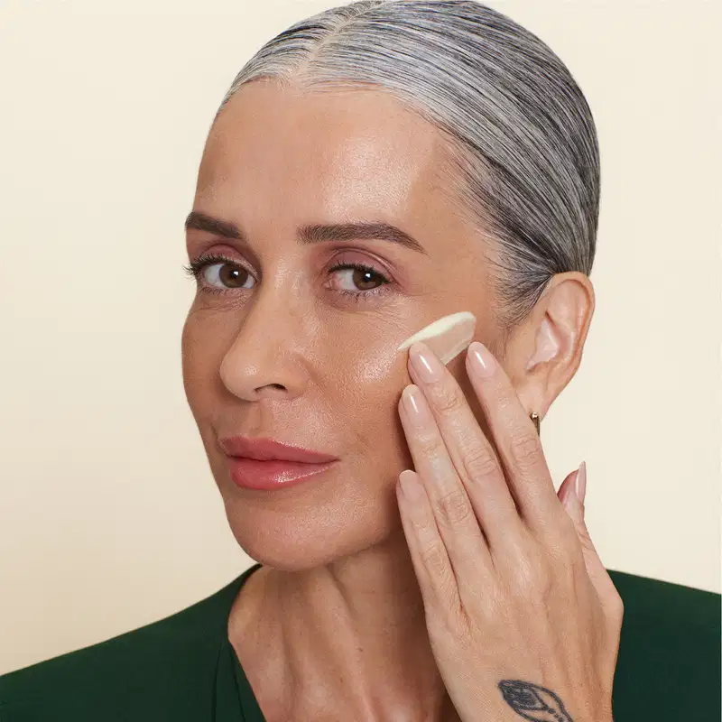 Imagem de uma mulher madura de cabelos grisalhos, com expressão séria, tocando o rosto com a mão e espalhando o creme facial. Ela usa um casaco verde e está em um fundo bege claro.