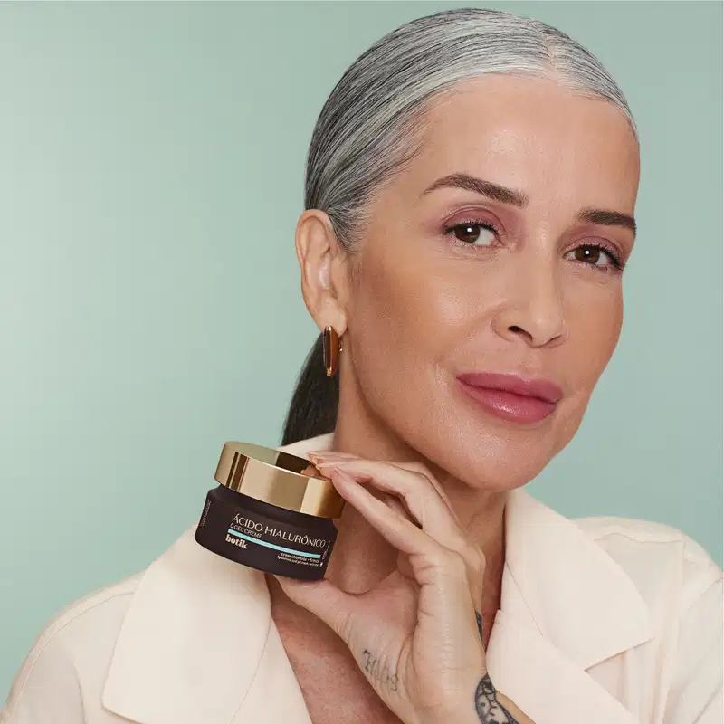 Imagem de uma mulher madura com cabelos grisalhos segura um frasco de creme facial da marca Botik em um fundo verde claro, com uma expressão séria e concentrada. O pote de creme é na cor vinho e possui tampa dourada.