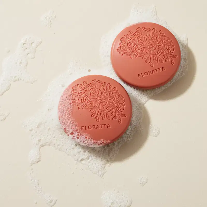 IImagem de dois sabonetes  redondos na cor rosa da marca Floratta Red em superfície branca molhada com espuma ao redor dos produtos.