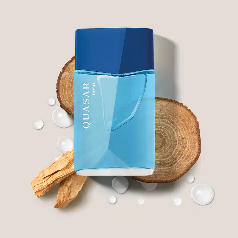 Imagem de um frasco de desodorante colônia da marca Quasar Rush em azul-claro com tampa azul royaç, sobre superfície de madeira com pedaços de casca e gotas de água.