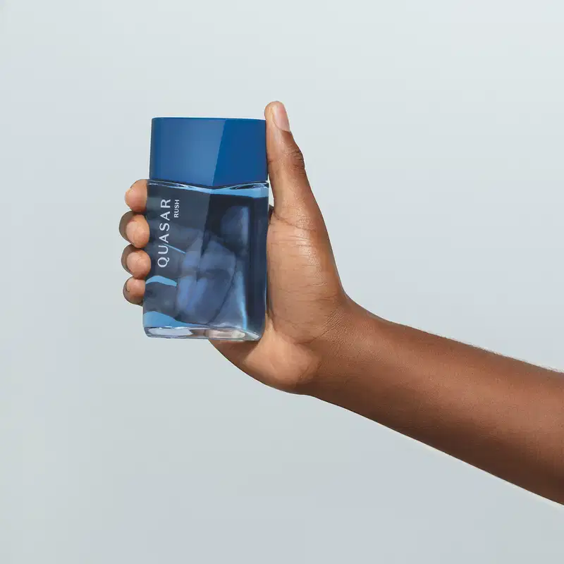 Imagem de um frasco em tons de azul de desodorante colônia Quasar Rush sendo segurado por mão masculina contra fundo branco liso.