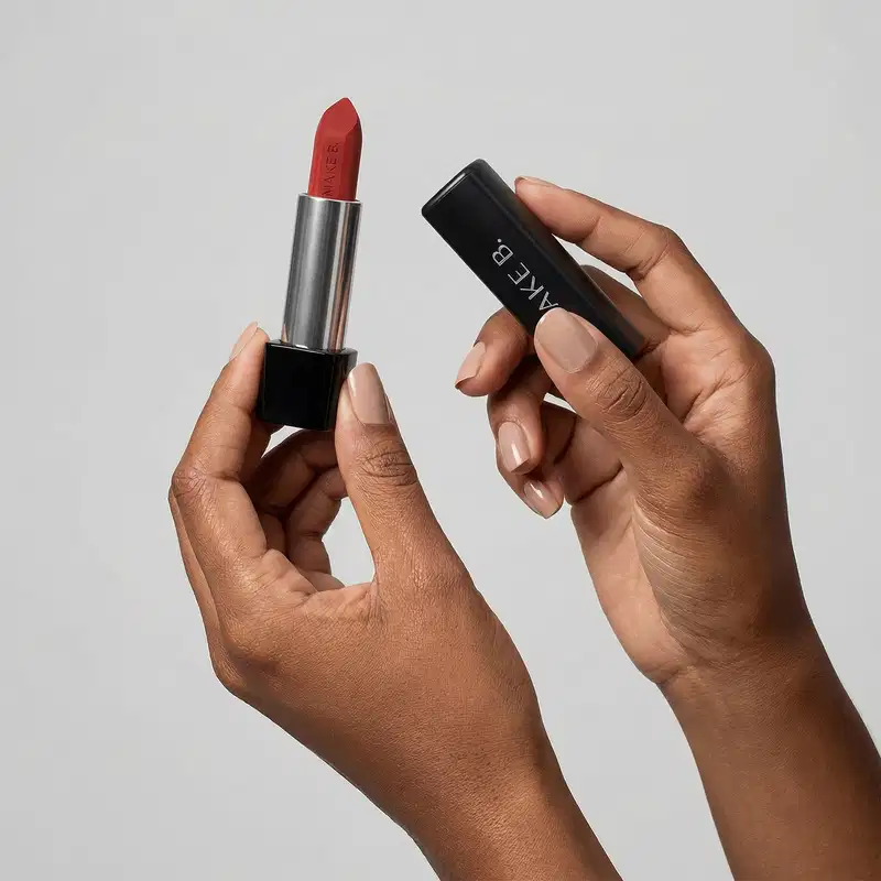 Mãos femininas segurando a tampa preta de um batom da marca Make B. na mão direita e a base do batom com a bala aparente na mão esquerda, em um ambiente minimalista e claro.