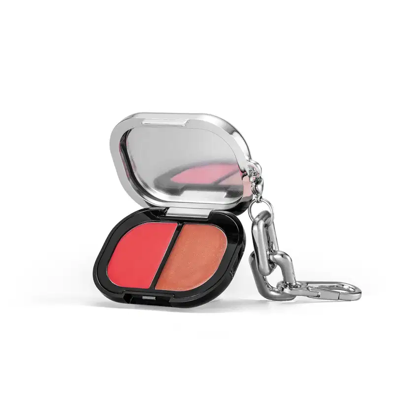 A imagem mostra o estojo do blush compacto aberta, apresentando as duas cores do produto em uma embalagem preta e prata. Ele possui um pingente metálico em formato de gancho preso à tampa.