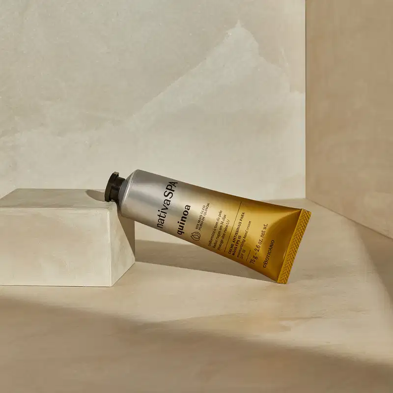 Imagem do tubo de creme de hidratação para as mãos Nativa SPA Quinoa deitado em superfície clara com sombras, em ambiente minimalista com parede clara e textura suave. A embalagem é prateada e dourada, possui tampa preta e informações em letras pretas.
