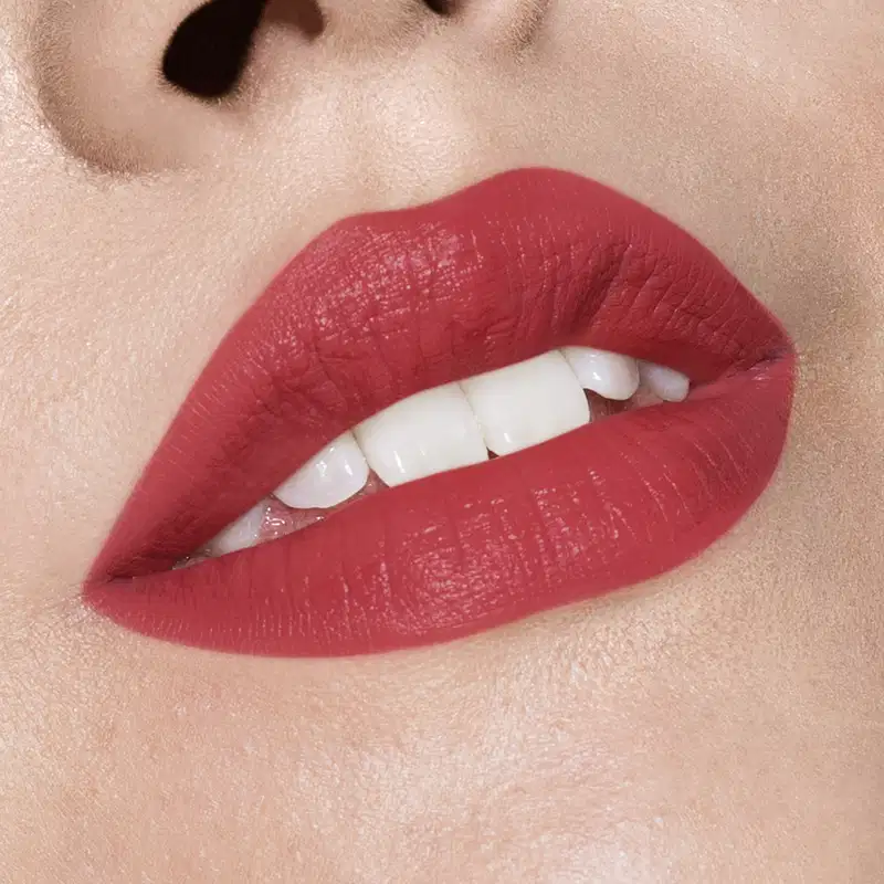 Imagem de lábios femininos em close-up com batom vermelho intenso. A boca tem um leve sorriso e está entreaberta, mostrando parcialmente os dentes brancos.