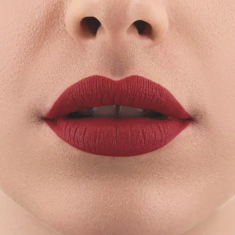 Imagem de um rosto feminino em close-up, evidenciando os lábios com o batom líquido Make B. na cor vermelho intenso. A boca está entreaberta mostrando parcialmente os dentes brancos.