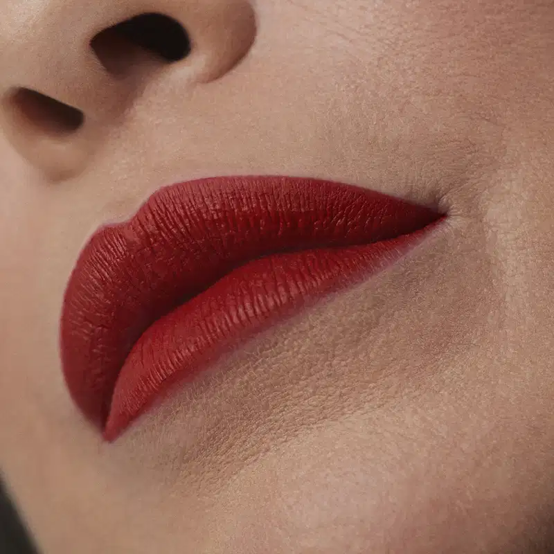 Imagem de um rosto feminino em close-up, evidenciando os lábios com o batom líquido Make B. em tom vermelho bem forte. A boca está fechada com um semblante sério.