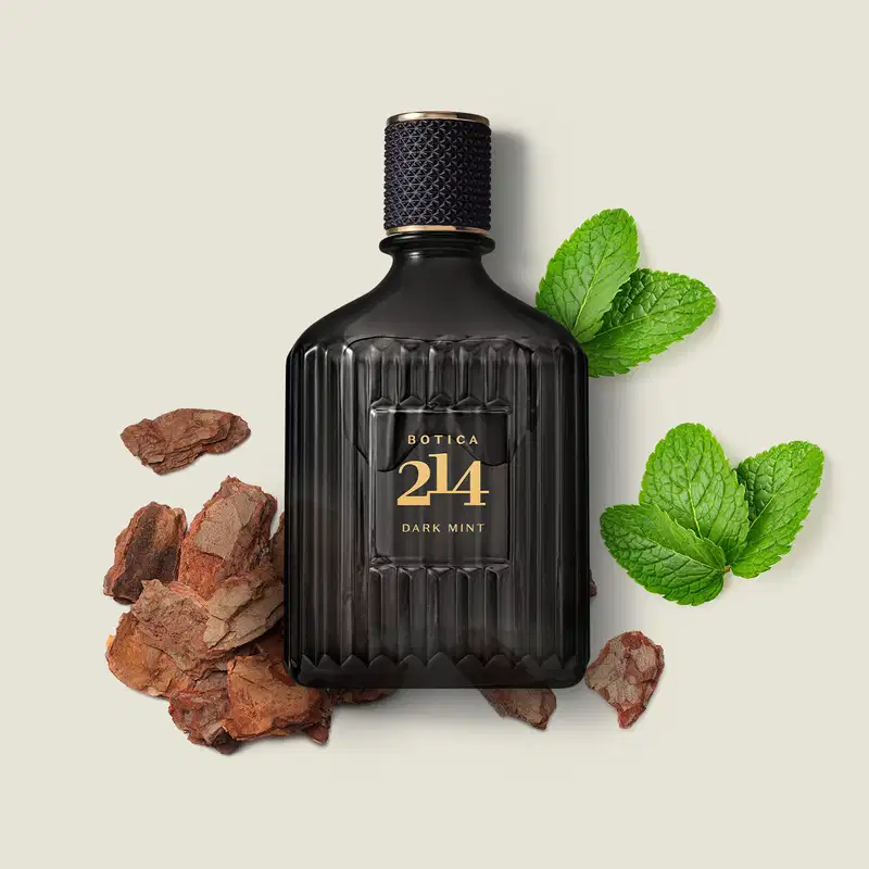 Frasco de vidro preto canelado com rótulo dourado e tampa texturizada de eau de parfum da marca Botica 214 Dark Mint cercado por lascas de madeira e folhas de menta sobre fundo claro.