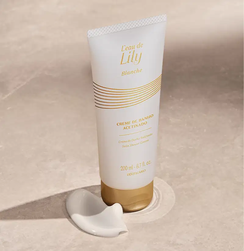 Imagem de um frasco branco com tampa e rótulo dourados de creme de banho acetinado da linha L'eau de Lily Blanche em fundo bege com sombras e pequena quantidade do produto ao lado da embalagem.