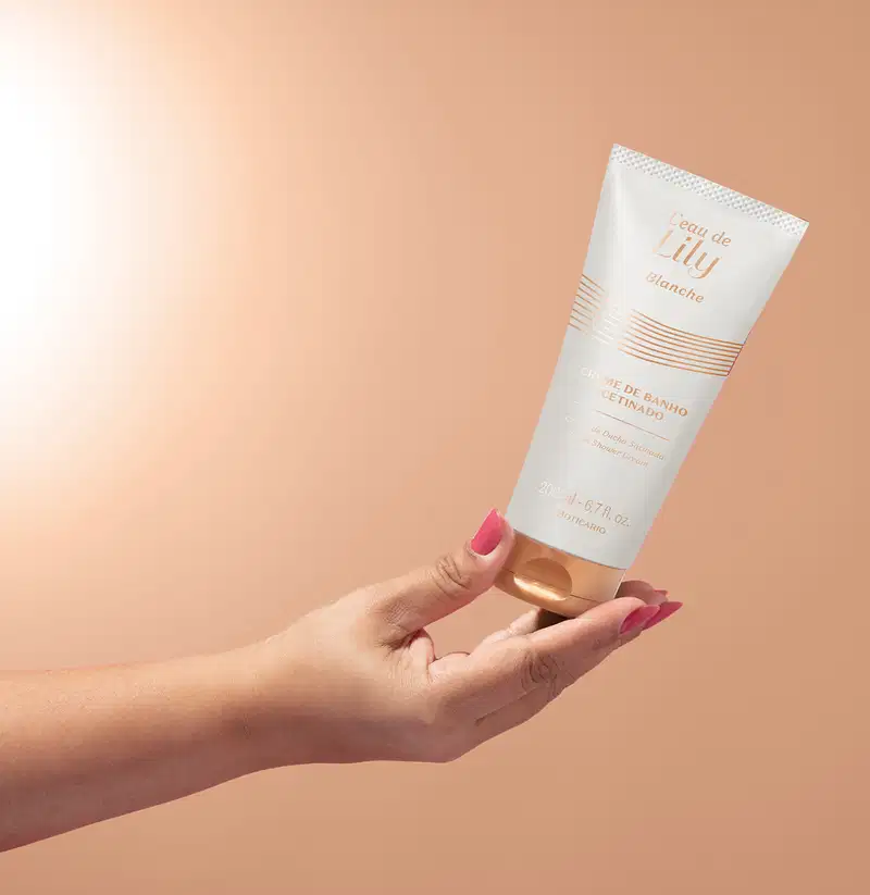Imagem de uma mão segurando um frasco branco com tampa e rótulo dourados de creme de banho acetinado da linha L'eau de Lily Blanche em fundo com degradê de branco e rosa.