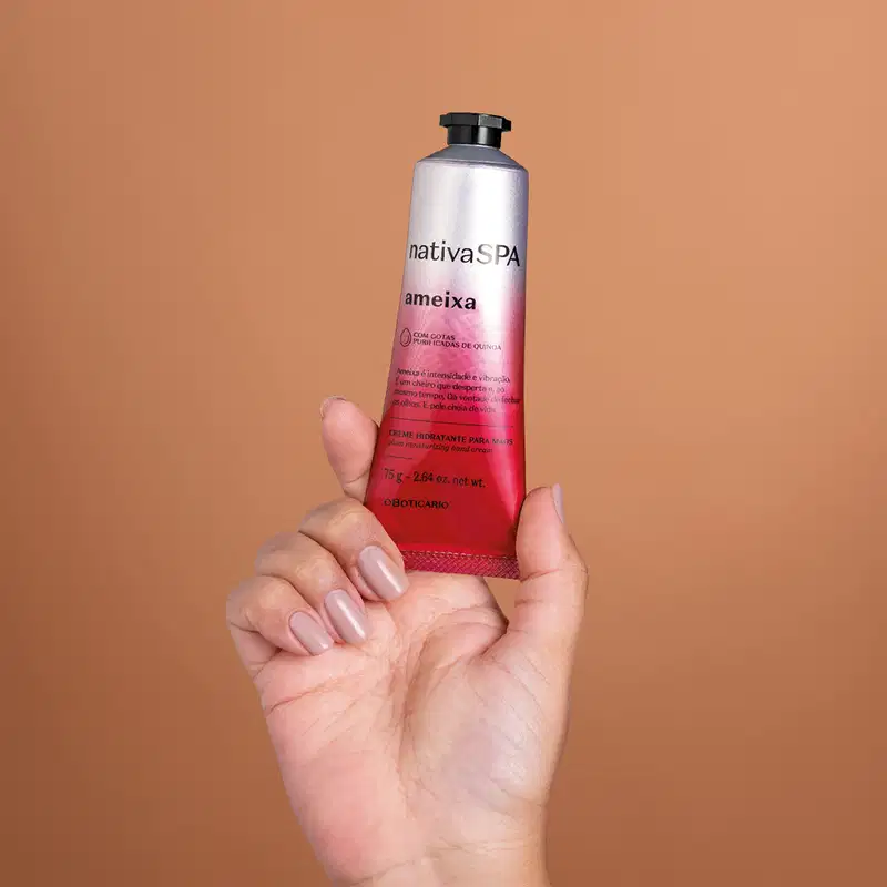 Imagem de uma mão feminina com unhas longas segurando um tubo de creme hidratante para as mãos Nativa SPA Ameixa. A bisnaga é na cor prata e rosa, possui tampa preta e informações sobre o produto estão impressas em letras pretas.. O fundo é bege.
