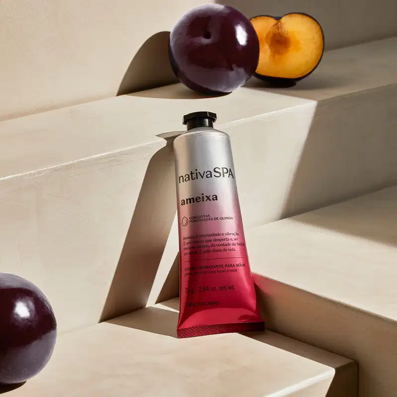 Imagem da embalagem prata e rosa do creme hidratante para as mãos Nativa SPA Ameixa sobre superfície clara, com ameixas roxas inteiras e cortadas ao meio ao redor, em ambiente minimalista e neutro.