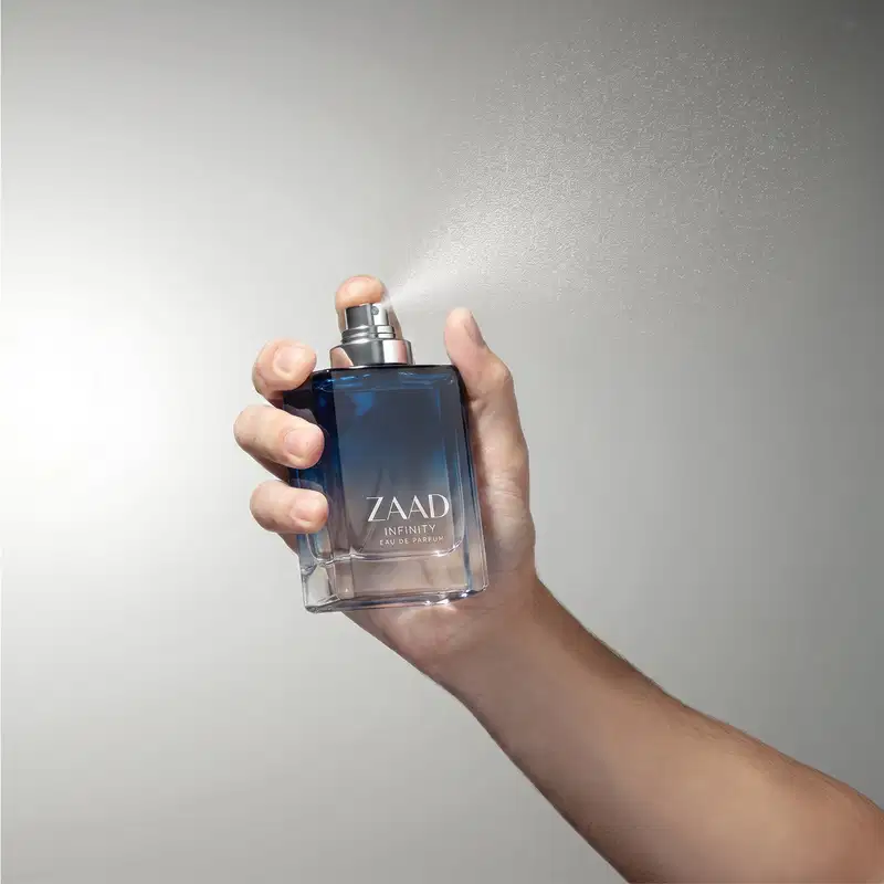 Frasco de item de perfumaria da marca Zaad Infinity em mãos contra fundo branco, com névoa de pulverização saindo do borrifador.