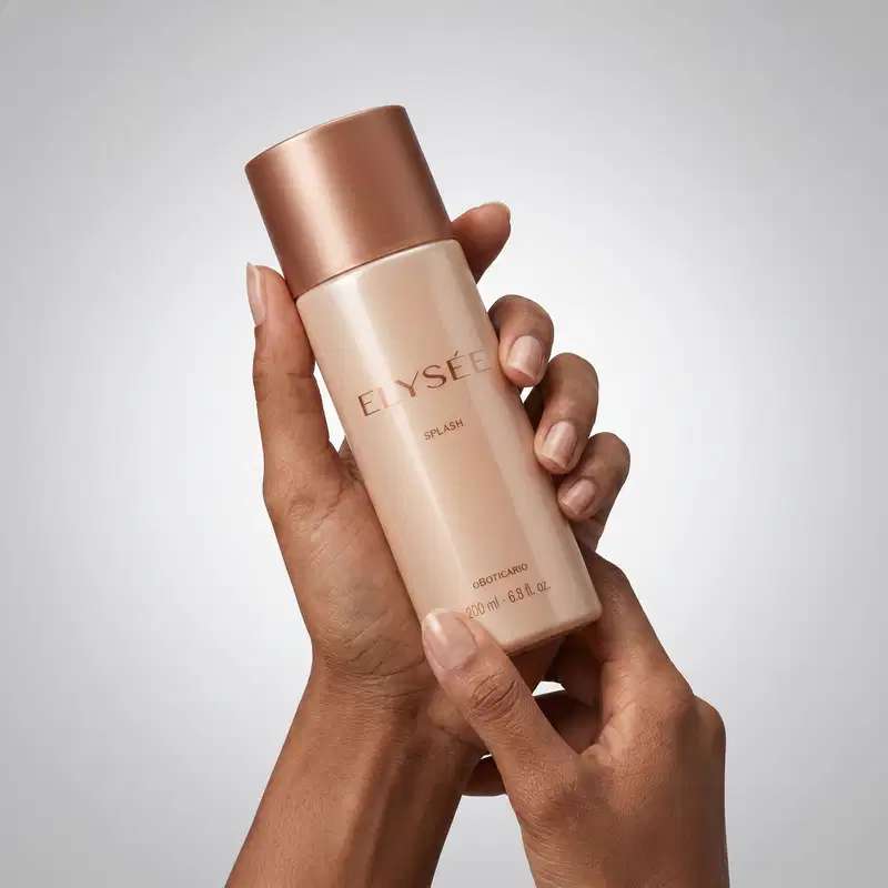 Frasco de item de perfumaria da marca Elysée em tom rosado e tampa rosé gold, sendo seguro por mãos femininas de cor branca em fundo cinza clara.