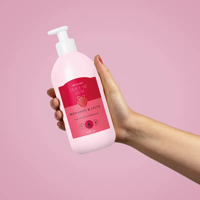 Imagem de uma embalagem branca de loção hidratante corporal Cuide-se Bem Morango e Leite com rótulo vermelho e rosa e imagem de morango, segura por mão feminina contra fundo rosa.