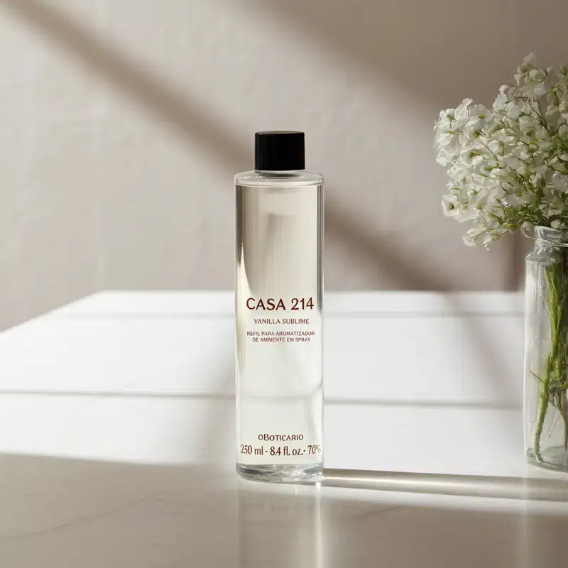 Imagem de um frasco refil com a fragrância Vanilla Sublime para difusor de ambiente da marca Casa 214 em embalagem transparente e tampa preta, posicionado sobre superfície clara com vaso de flores brancas ao fundo.