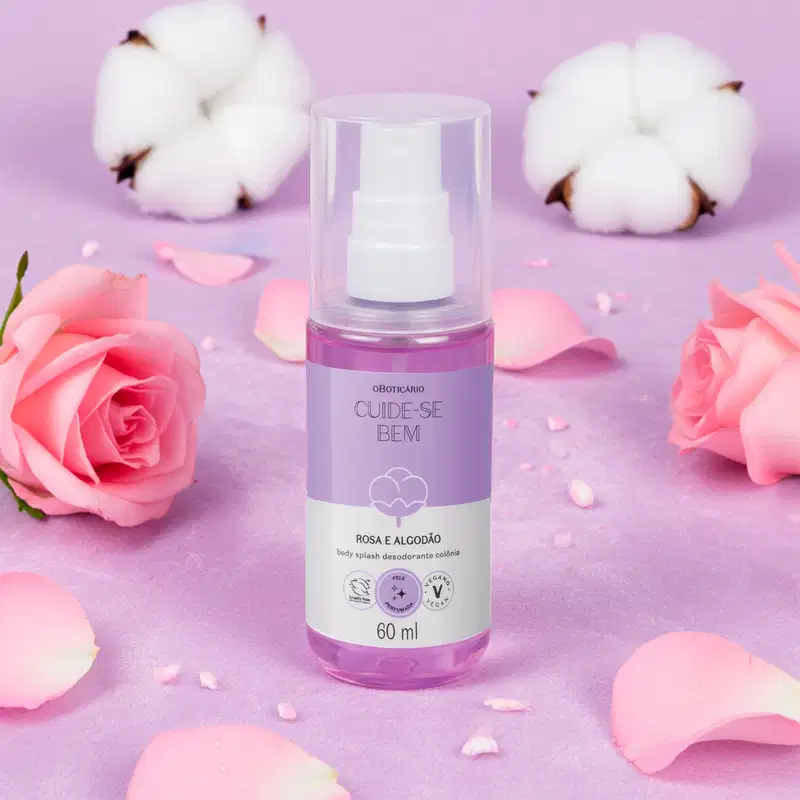 Imagem de um frasco cilíndrico transparente de body splash da marca Cuide-se Bem Rosa e Algodão com líquido rosa, rótulo bicolor lilás e branco com ilustração de um floco de algodão em cenário de fundo roxo com rosas e bolas de algodão espalhadas.