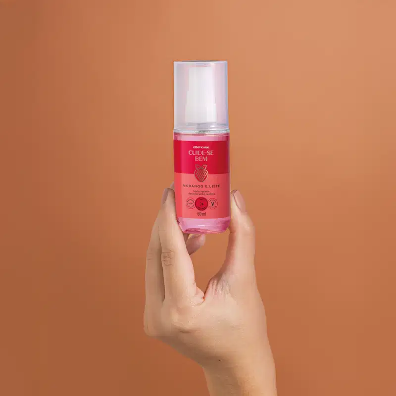 Imagem de uma mão feminina segurando um vidro transparente com líquido rosa, borrifador branco e tampa transparente contendo body splash Cuide-se Bem Morango e Leite. O fundo é rosa vibrante.