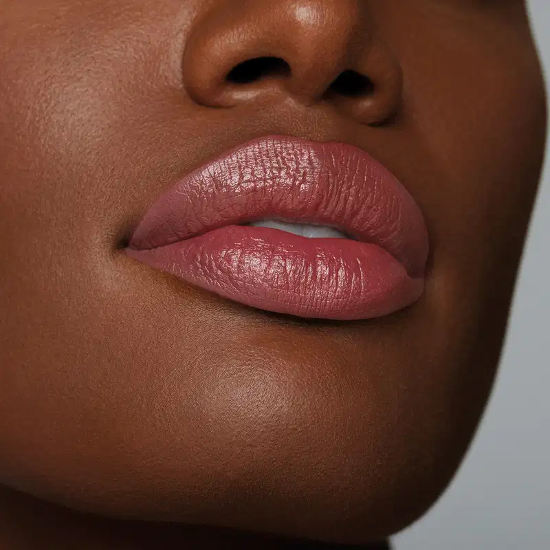Boca feminina de mulher negra em close mostrando queixo e um pedaço do nariz, com lábios levemente entreaberto, revelando um pouco dos dentes frontais brancos e lábios com batom rosado sobre eles.